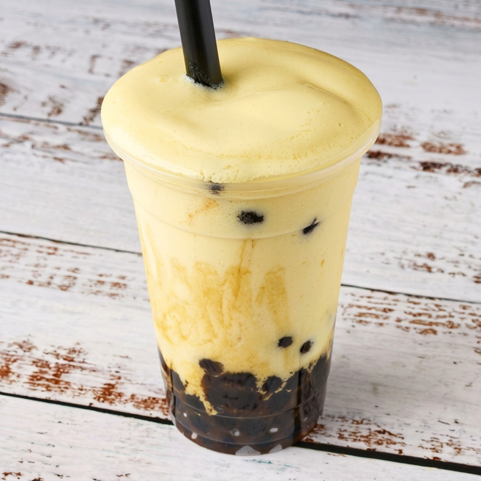 Yellow mango smoothie with black tapioca pearls in a clear cup, topped with foam and a straw on a rustic white wooden table.